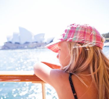 Young blond girl wearing a hat, leaning on a ferry railing, and looking at sites with Sydney Opera House in the background