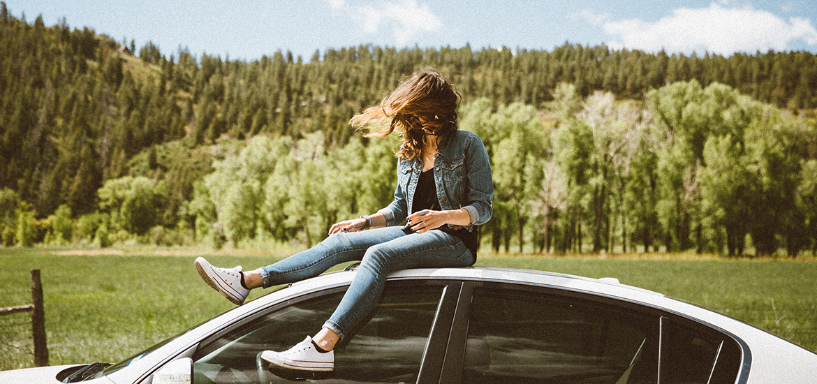 Woman wearing stylish jeans, jean jacket, a black shirt, & white Converse shoes sitting atop a car